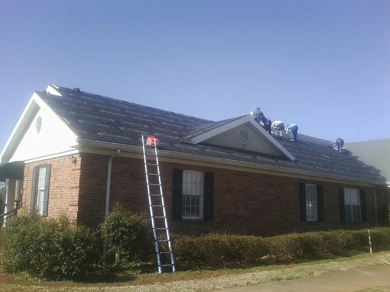 Crew tearing shingles off a brick ranch roof with a ladder set — Metal Roof Repair work by our crew in Kenly