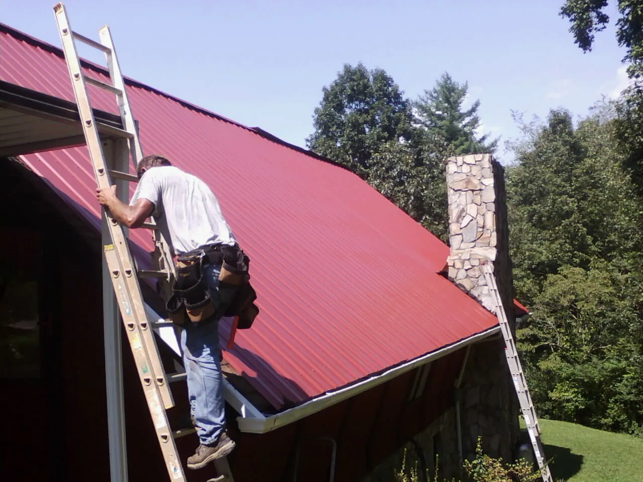 Roofer on a ladder beside a red metal roof and stone chimney — Soffit & Fascia Repair by Roofing Brothers in Kenly